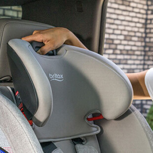 A woman is standing by the open back door of a car, adjusting a forward-facing car seat that is installed in the back seat. 