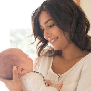 Smiling parent cradles a newborn near a bright window.