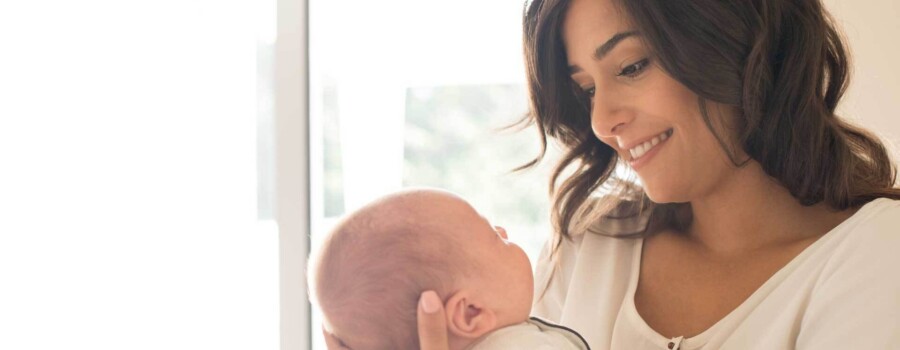 Smiling parent cradles a newborn near a bright window.