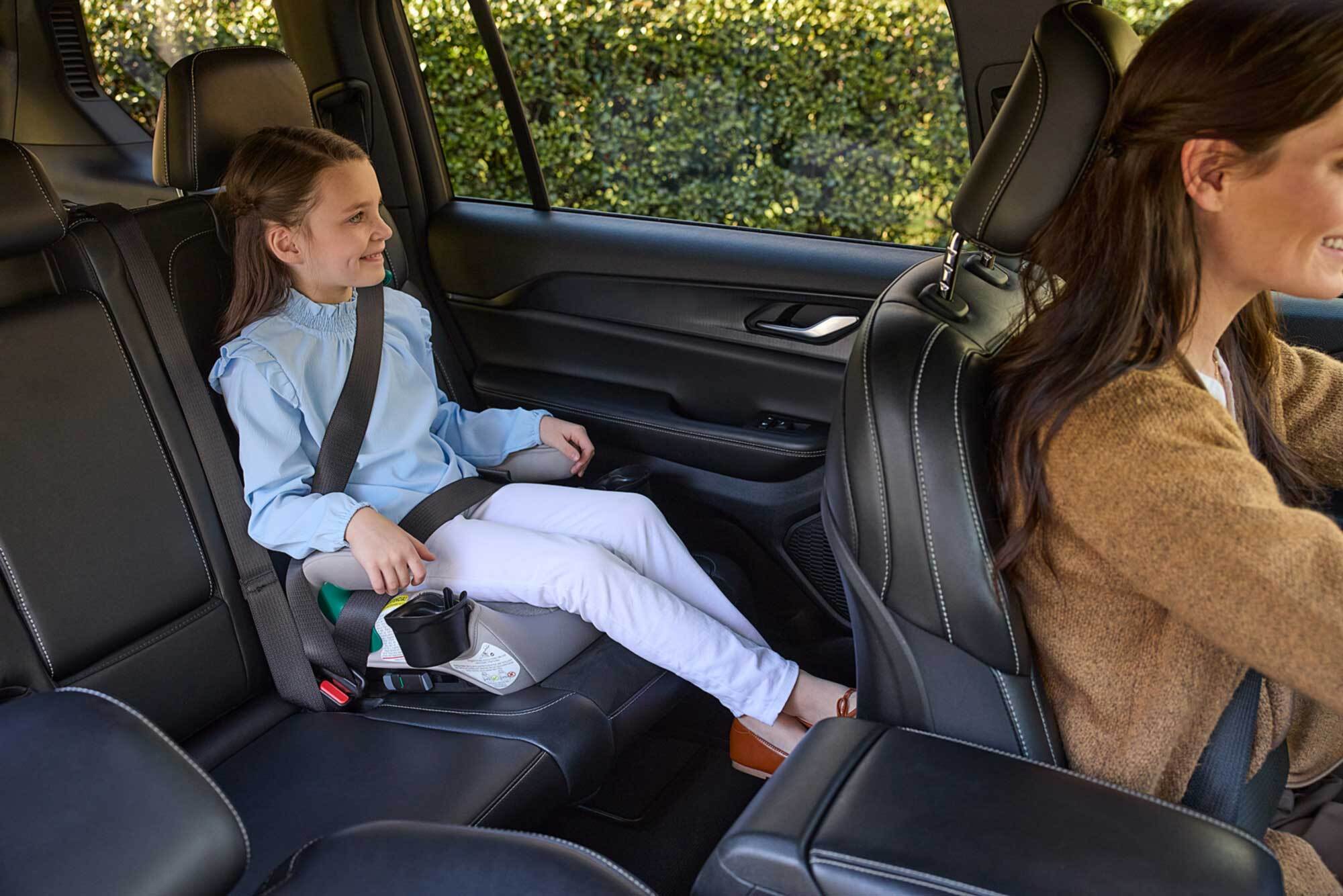 A child sitting in a booster car seat in the back seat of a vehicle, buckled in and looking out the window.