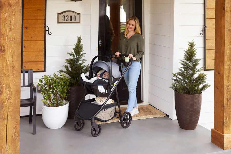 A woman standing on a porch in front of a door. She is pushing a baby in a stroller.