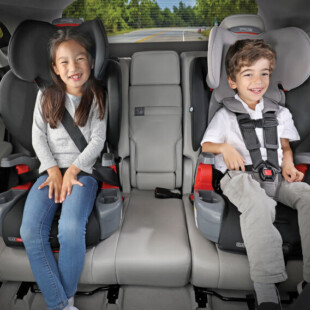 Two smiling children in the back seat of a vehicle. One is in a booster, and the other is in a car seat with a harness.