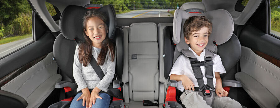Two smiling children in the back seat of a vehicle. One is in a booster, and the other is in a car seat with a harness.