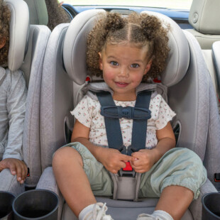 Three children of varying ages buckled into car seats that are lined up 3-across in the back seat of a car.