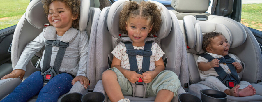 Three children of varying ages buckled into car seats that are lined up 3-across in the back seat of a car.
