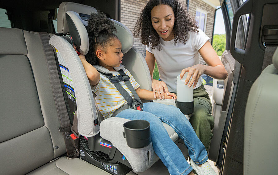 A young child in a forward-facing car seat in the back of a vehicle. An adult is placing a bottle into the car seat cup