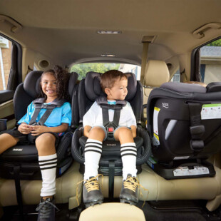 Two children safely buckled in forward-facing car seats next to a third rear-facing car seat.