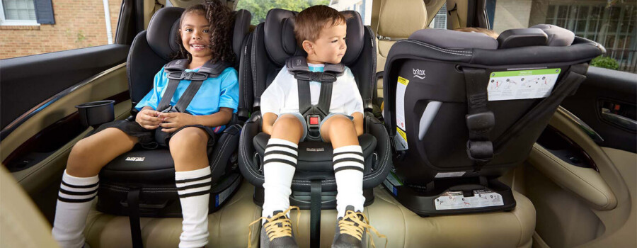 Two children safely buckled in forward-facing car seats next to a third rear-facing car seat.