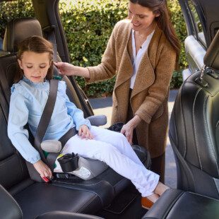 A woman adjusts the vehicle seat belt as a child buckles up in a booster car seat.
