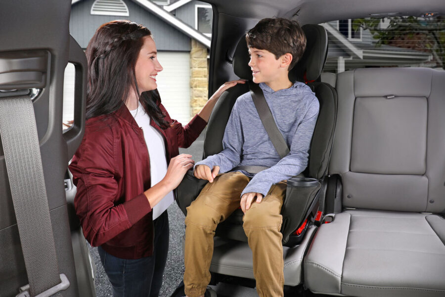 A woman standing outside of a vehicle with an open back door, smiling at a child who is sitting in the car in a booster seat.