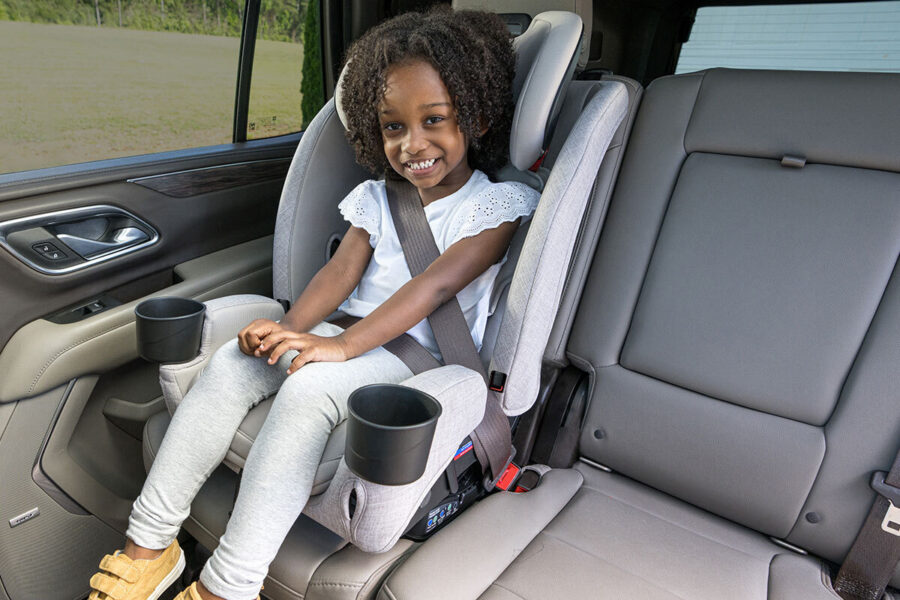 A smiling child buckled up in the back seat of a car, sitting in a high-back booster seat. 