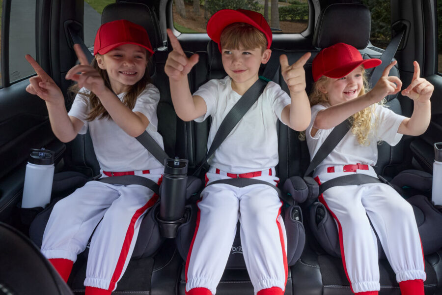 Three children wearing baseball uniforms, sitting in backless booster car seats in the back seat of a vehicle.
