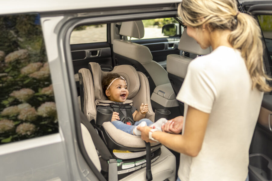 A woman standing by the open rear door of a vehicle, interacting with an infant in a rotating car seat.