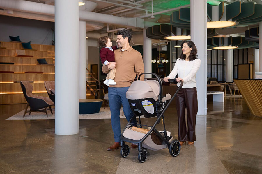 A man holding a toddler in his arms while a woman pushes a stroller with an infant car seat attached, inside of a building.