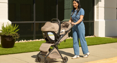 A woman pushing a stroller with an infant car seat attached outside on a sunny day.