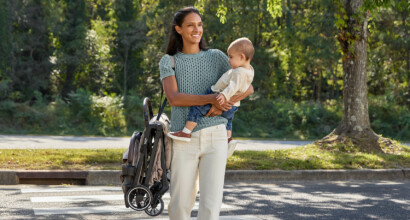 A woman carrying a toddler and walking in a crosswalk while carrying a folded stroller with a strap over her shoulder.