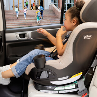 A child in a car seat, looking out of the car window at other children entering a school.
