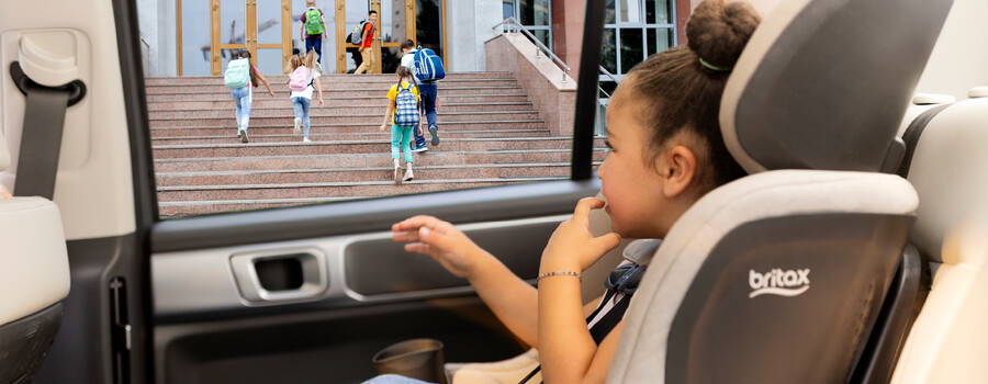 A child in a car seat, looking out of the car window at other children entering a school.