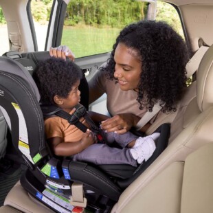 Someone is leaning into the back seat of a vehicle and smiling at a young child, who is harnessed into a rear-facing car seat. 