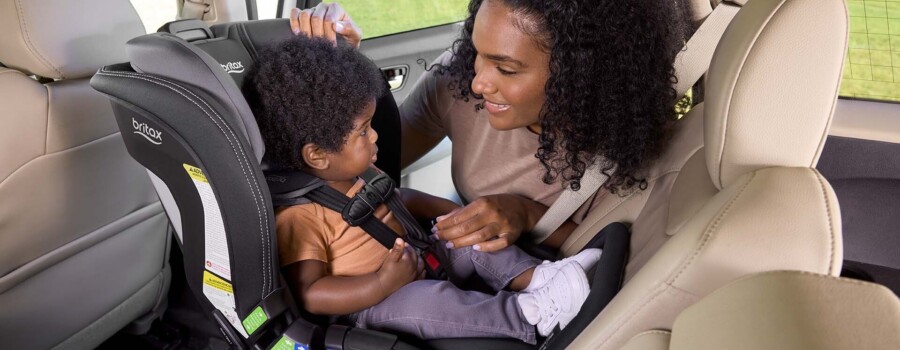 Someone is leaning into the back seat of a vehicle and smiling at a young child, who is harnessed into a rear-facing car seat. 