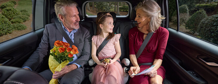 Two nicely dressed adults and a child sitting in the back seat of a vehicle. The child is in a booster seat in the middle.