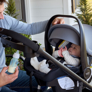 A man sitting and holding a baby bottle while watching a baby in a travel system stroller.