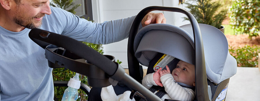 A man sitting and holding a baby bottle while watching a baby in a travel system stroller.
