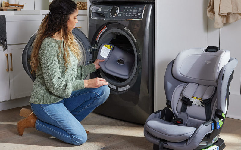 Woman kneeling next to a child car seat and placing an item into a washing machine.