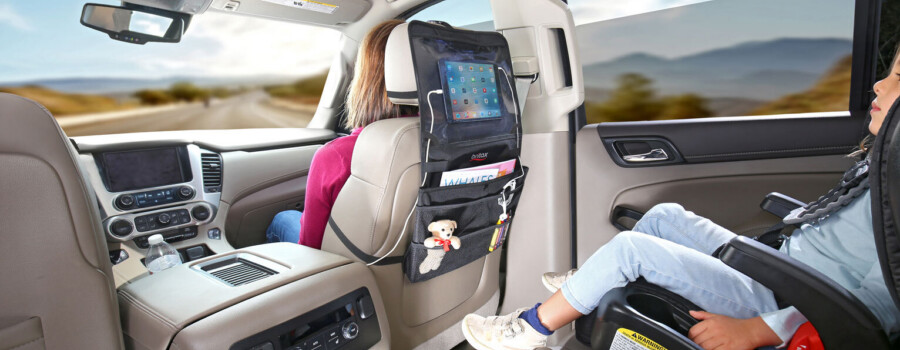 A child in a car seat in the back of a vehicle looking at a tablet. The tablet is inside of a vehicle seatback organizer.