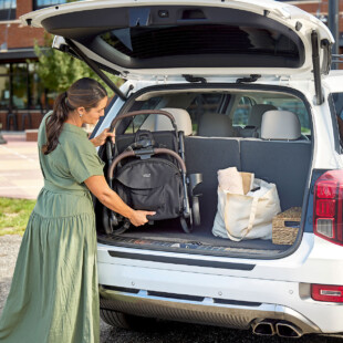 A woman putting a baby stroller into the trunk of a car alongside a bag and a small basket.