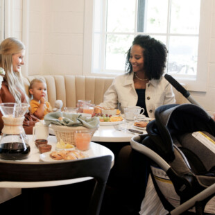 A group of women with two babies, sitting around a table set with various breakfast foods.