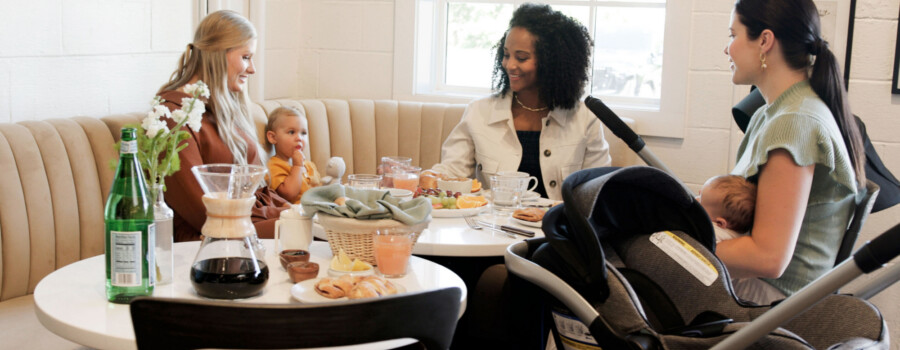 A group of women with two babies, sitting around a table set with various breakfast foods.