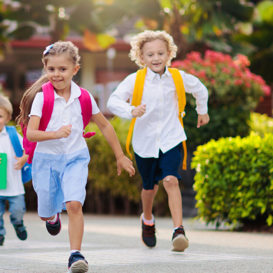 Two children wearing backpacks and running on the sidewalk.