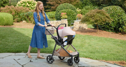 A woman pushing a stroller with an infant car seat attached, walking along an outdoor pathway on a sunny day.