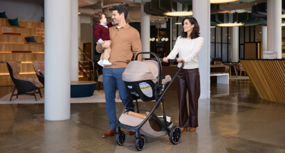 A family walking together inside a modern building. The man is carrying a toddler, and the woman is pushing a travel system stroller.