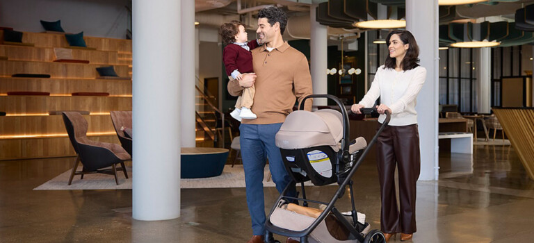 A family walking together inside a modern building. The man is carrying a toddler, and the woman is pushing a travel system stroller.