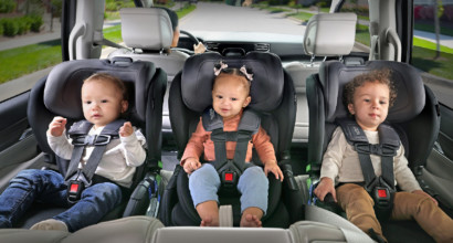 Three children of varying age in rear-facing car seats lined up three-across in the back seat of a car.