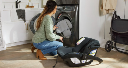 A woman placing some padding into a washing machine. There is a car seat and a stroller on the floor next to her.