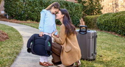 A child in front of a house, holding a booster car seat and smiling at a woman, who is kneeling next to her.