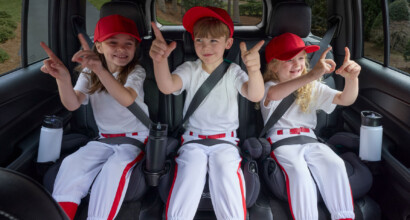 Three children in baseball outfits, sitting in the back row of a car in booster seats, smiling and making playful gestures.